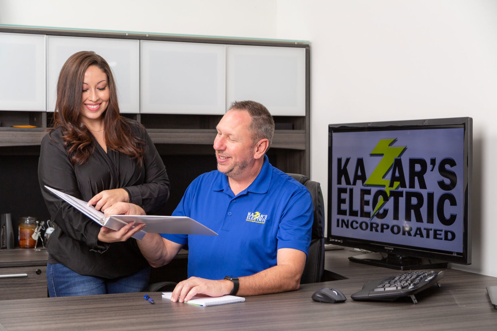 Man and woman discussing project details at Kazar's Electric office, with logo visible on computer screen, showcasing teamwork and professionalism in electrical services.