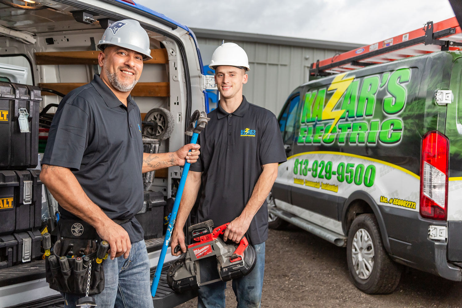 Two Kazar's Electric technicians smiling in front of their service van, one holding a pipe wrench and the other a power tool, showcasing their expertise in electrical services for commercial and residential needs.