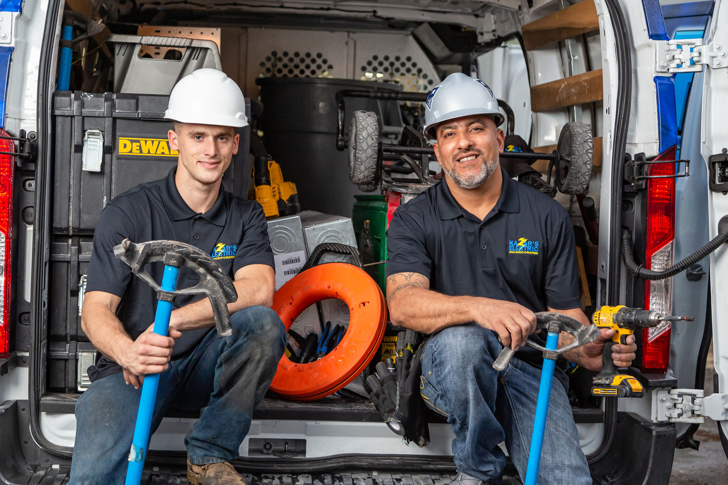 Two Kazar's Electric technicians in hard hats, seated in a work van filled with electrical tools and equipment, showcasing their commitment to providing quality electrical services.
