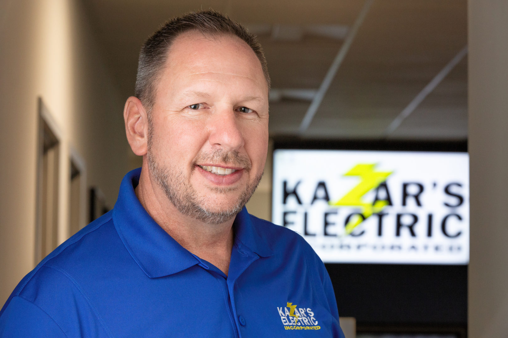 Man in blue polo shirt with Kazar's Electric logo, smiling in front of the company's logo displayed in the background, representing trusted electrical services in Tampa.