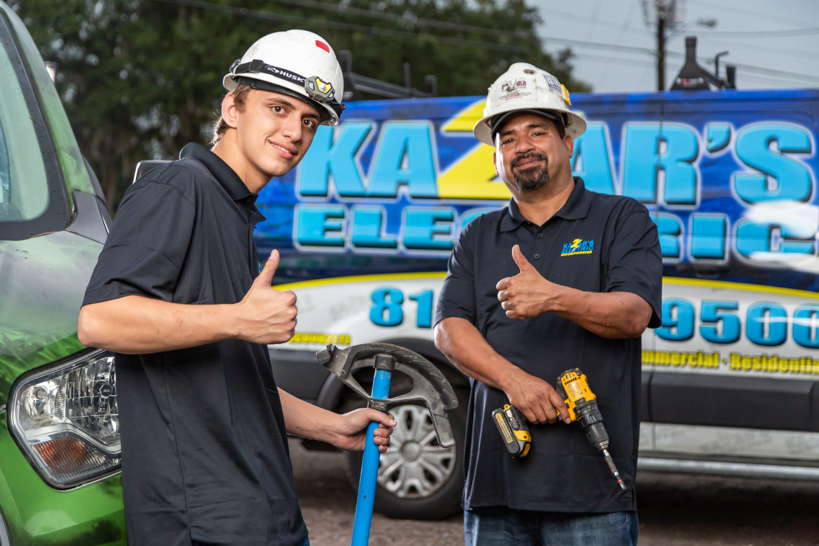 Two Kazar's Electric technicians giving thumbs up, one holding a tool and the other a drill, with a branded service vehicle in the background, promoting 24/7 emergency electrical services in Tampa.