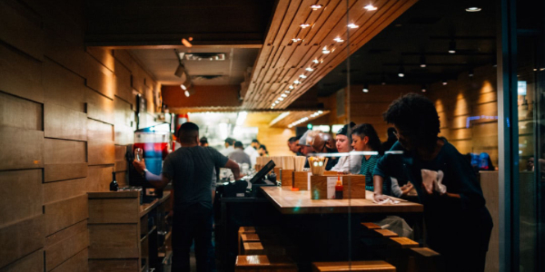 Interior of a busy restaurant showcasing commercial lighting design, with staff serving customers at a wooden bar and overhead lights creating an inviting atmosphere.
