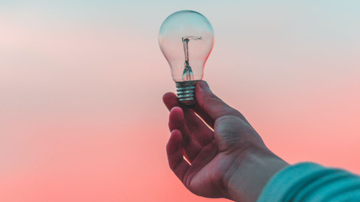 Hand holding a light bulb against a colorful sky, symbolizing energy conservation and electrical services.