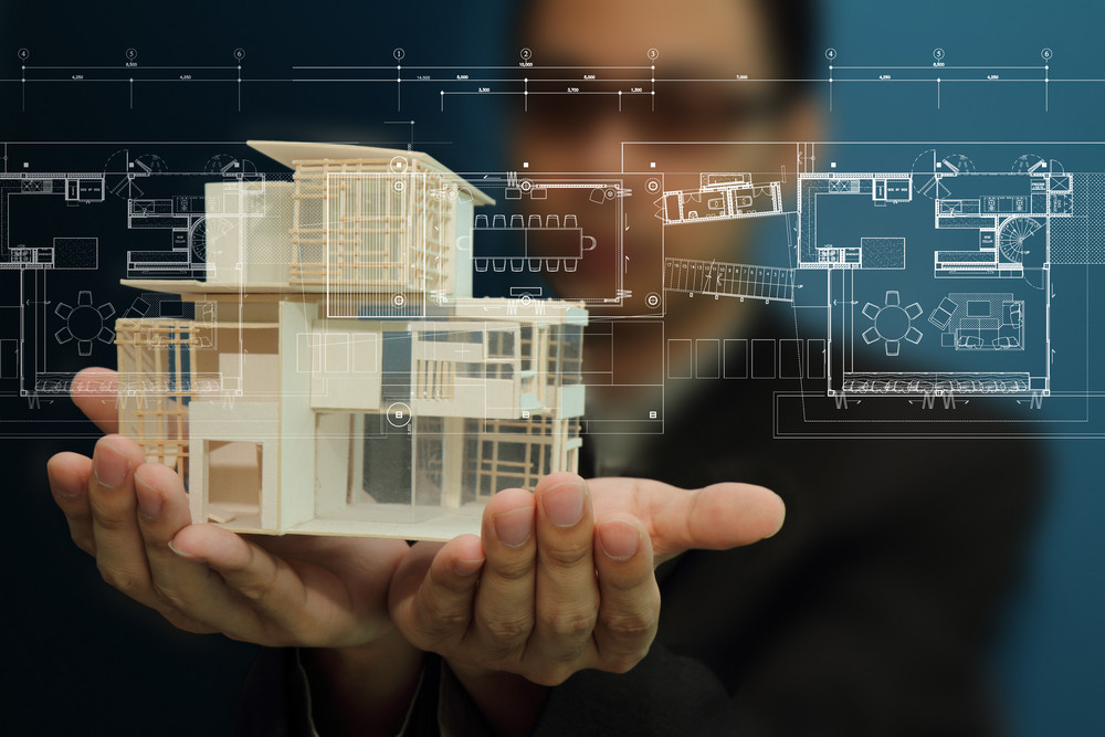 Person holding a model house with architectural blueprints overlay, illustrating electrical planning for new home construction.