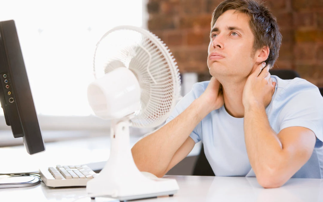 Man sitting at a desk with a computer and a small fan, looking upward with a thoughtful expression, illustrating the need for effective cooling solutions like ceiling fans to enhance comfort and energy efficiency in homes.